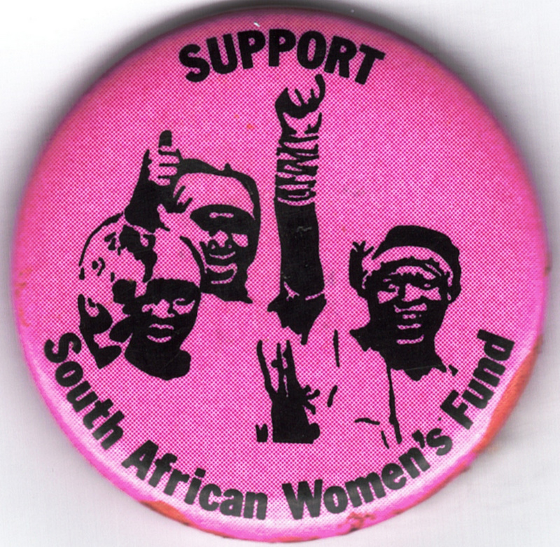 Round, pink metal anti-apartheid button. Three women who are smiling and raising an arm are at the center of the button, with text around the top and bottom. The image and text are in black. Size: 1-1/2 inches across.

